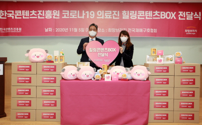 붙임. 힐링 콘텐츠 BOX 전달식 현장사진 사진 1. 5일 서울 마포구에 위치한 희망브리지에서 코로나19의 장기화 상황 속 방역현장 최전선을 지키는 의료진을 위해 콘진원이 마련한 응원 물품인 '힐링 콘텐츠 BOX' 전달식이 진행되었다.(좌측 콘진원 김영준 원장, 우측 희망브리지 전국재해구호협회 정서윤 본부장)